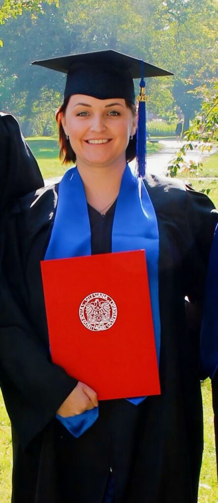 Iva Bártková, lektorka angličtiny a majitelka jazykové školy Top English, stojí v zahradě, má na sobě vysokoškolský graduační hábit, graduační čepičku a drží v ruce svůj červený diplom.